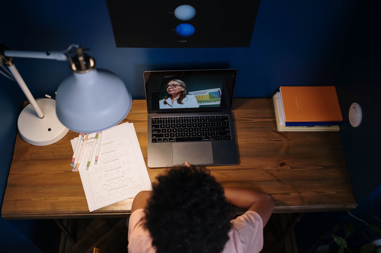 gallery-1 A student attends a virtual class, studying at a wooden desk filled with educational materials.